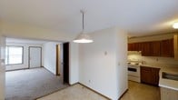 This single-floor residential space offers a practical and functional layout designed for comfortable everyday living. The open-concept design connects the kitchen and living areas seamlessly, creating a cohesive flow throughout the unit. The kitchen is equipped with a white electric range, a ventilation hood, and warm wood-toned cabinetry, providing ample storage and a classic, understated aesthetic. A pendant light with a chain mount hangs at the transition between the kitchen and the adjoining living space, serving as a subtle focal point and providing direct task lighting. The living area features neutral carpeting that extends through the space, contributing to a warm and quiet ambiance. Crisp white walls and white trim work throughout give the interior a clean, bright appearance. A hallway visible from the main living area leads to additional rooms, suggesting a well-organized floor plan. Recessed doorways and standard-height ceilings maintain a cozy and intimate scale. Overall, the space presents a straightforward residential environment with classic finishes that offer a solid foundation for personalization.