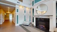 This refined communal space presents a grand, welcoming atmosphere defined by soaring double-height ceilings, crisp white architectural millwork, and vibrant teal accent panels that lend a distinctive, polished character throughout. The centerpiece of the main lounge area is a striking fireplace framed by an elaborate white surround, topped with a large circular mirror that amplifies the sense of openness. Flanking built-in display niches with pendant lighting showcase decorative objects, adding warmth and visual interest. Comfortable upholstered armchairs and a soft area rug create an inviting seating arrangement around the hearth. Tall decorative columns with teal inlays divide the space, framing the transition into a wide, well-lit corridor. The hallway features warm tile flooring, white wainscoting, and recessed lighting, with framed artwork adorning the walls and clear signage guiding visitors. A secondary seating area near the entry offers a sofa and accent chair alongside large glass doors that invite natural light inside. Throughout, the interplay of warm neutrals, teal accents, and bright white trim creates a cohesive, elegant ambiance suited to a professional or residential community building.