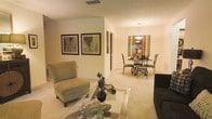 This single-floor residence offers a comfortable and well-appointed living environment with a seamless, open layout that connects the main living and dining areas. The living space is anchored by plush carpeted flooring and features a glass-topped coffee table, a cushioned accent chair, and wall art that adds a refined, curated touch to the warm, neutral-toned walls. Natural flow carries through to the dining area, where a round table surrounded by elegant wrought iron chairs sits beneath a classic pendant chandelier, creating an intimate setting for meals. A decorative mirror and warm ambient lighting enhance the dining nook's welcoming character. The open sightlines between rooms contribute to an airy, spacious feel throughout the home. Soft, neutral tones on the walls and flooring create a cohesive palette that makes the space feel bright and inviting. The single-floor layout ensures easy accessibility and a practical flow between areas. Overall, the residence strikes a balance between functional design and understated elegance, making it a comfortable and visually appealing living environment.