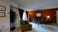 This single-floor residence centers around a comfortable and thoughtfully designed living room that serves as the focal point of the space. The room is distinguished by a bold terracotta-red accent wall, which adds warmth and depth to the overall aesthetic, complemented by neutral white ceilings and light-toned carpeting throughout.

The living area is generously proportioned, accommodating a full seating arrangement that includes a large sofa with decorative accent pillows and a matching armchair, both upholstered in earthy brown tones. A wrought-iron coffee table anchors the seating area, adding a classic decorative touch. A tall floor lamp provides ambient lighting, while a framed mirror on the accent wall enhances the sense of space.

Natural light enters through multiple windows dressed in rich burgundy curtains, including a sliding glass door that opens to an outdoor balcony or patio, where outdoor seating is visible. A separate dining nook near a window accommodates a small table and chairs. Decorative indoor plants are positioned throughout, adding a natural, lived-in quality. The ceiling fan and visible wall-mounted HVAC unit contribute to the unit's practical amenities.