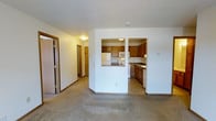 This single-floor residential unit offers a functional and well-organized layout designed for comfortable everyday living. The living area features neutral carpeting and smooth white walls, creating a clean and inviting atmosphere. A textured ceiling adds a classic finishing touch throughout the space.

The kitchen is thoughtfully positioned adjacent to the living area, separated by a half-wall with a pass-through opening that maintains a sense of connection between the two spaces. The kitchen is equipped with warm honey-toned wood cabinetry offering ample storage, white appliances including a refrigerator, range, built-in microwave, and dishwasher, along with a countertop sink. The kitchen floor features resilient vinyl or tile flooring, providing a practical contrast to the carpeted living area.

A hallway extends from the main living space, lined with doors that lead to additional rooms, including what appears to be a closet or storage area, suggesting well-distributed storage throughout the unit. The corridor is illuminated by a ceiling-mounted light fixture, ensuring the space remains bright and navigable. Overall, the unit presents a tidy, move-in-ready environment with a straightforward and efficient floor plan.
