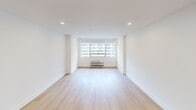 This thoughtfully designed single-floor unit at 1512 offers a clean, modern aesthetic with high-quality finishes throughout its two principal spaces. The main living area is an open, airy room characterized by crisp white walls, light-toned hardwood flooring, and recessed ceiling lighting that creates a bright and welcoming atmosphere. A wide, multi-pane window with roller shades anchors the far wall, flooding the space with natural light and offering views of the surrounding urban skyline. A wall-mounted PTAC unit ensures year-round climate control. The open layout connects seamlessly to a compact kitchenette tucked into a alcove, which features light wood cabinetry, a stainless steel refrigerator, an under-cabinet sink, and recessed lighting — providing efficient functionality within a minimal footprint. The bathroom is a standout space, showcasing a refined, contemporary design with large-format white marble-look countertops, a wood-toned vanity base, a sleek black-framed mirror, and matte black hardware and fixtures throughout. A glass-enclosed shower with marble-style tile walls adds a polished, upscale touch. The consistent use of neutral tones and modern materials throughout the unit creates a cohesive, sophisticated living environment.