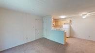 This single-floor residential unit offers a functional and efficient open-concept layout designed to maximize usability. The living area is finished with neutral wall-to-wall carpeting that adds warmth underfoot and a clean, understated look throughout. White walls and a flush ceiling light fixture create a bright, welcoming atmosphere. The kitchen is positioned adjacent to the living space and is separated by a peninsula counter, which provides both additional workspace and an informal bar or dining area. Light wood cabinetry lines the kitchen walls, offering ample storage, and is complemented by a white refrigerator and a stainless-style sink. The kitchen flooring transitions to a smooth hard surface, practical for food preparation areas. Two interior doors are visible near the entry area, suggesting access to closet or utility space, as well as additional rooms. The overall design is straightforward and practical, featuring a cohesive neutral color palette of whites and soft beiges throughout. The space is well-suited for everyday living, with a layout that keeps the kitchen and living areas visually connected while maintaining a sense of separation through the peninsula counter design.