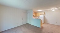 This single-floor residential unit offers a functional and efficient open-concept layout designed to maximize usability. The living area is finished with neutral wall-to-wall carpeting that adds warmth underfoot and a clean, understated look throughout. White walls and a flush ceiling light fixture create a bright, welcoming atmosphere. The kitchen is positioned adjacent to the living space and is separated by a peninsula counter, which provides both additional workspace and an informal bar or dining area. Light wood cabinetry lines the kitchen walls, offering ample storage, and is complemented by a white refrigerator and a stainless-style sink. The kitchen flooring transitions to a smooth hard surface, practical for food preparation areas. Two interior doors are visible near the entry area, suggesting access to closet or utility space, as well as additional rooms. The overall design is straightforward and practical, featuring a cohesive neutral color palette of whites and soft beiges throughout. The space is well-suited for everyday living, with a layout that keeps the kitchen and living areas visually connected while maintaining a sense of separation through the peninsula counter design.