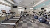 This fitness center offers a spacious, single-floor layout designed to accommodate a comprehensive range of workout routines. The main exercise area is outfitted with a robust selection of cardio equipment, including multiple treadmills, elliptical trainers, and a stair climber, all from reputable brands such as True Fitness and Precor. A dedicated section for additional strength and cross-training machines rounds out the equipment offerings.

The interior features a distinctive checkered carpet in shades of gray, adding a clean, modern aesthetic to the floor. Tray ceilings with recessed lighting and multiple ceiling fans ensure the space remains well-lit and comfortably ventilated throughout. Large windows fitted with white blinds line the walls, allowing generous natural light to fill the room while maintaining privacy.

Full-length mirrors are mounted along one wall, both enhancing the sense of openness and providing users with proper form feedback during workouts. Multiple wall-mounted flat-screen televisions are positioned throughout the space for entertainment during exercise sessions. The color scheme of soft gray-green walls accented with teal highlights creates a calm, motivating atmosphere. Clearly marked exits and accessible door hardware reflect thoughtful safety and accessibility considerations throughout the facility.