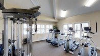 This fitness center offers a clean, purpose-built environment designed to support a well-rounded workout routine. The space is anchored by a multi-station cable weight machine positioned prominently, providing a range of strength training options. Alongside it, two treadmills and an elliptical trainer make up a solid cardio station, catering to various endurance and low-impact exercise preferences. The room is finished with durable, low-pile carpet flooring suited for a high-traffic fitness environment. Elevated ceilings with decorative beam detailing lend a sense of openness to the space, while flush-mounted overhead lighting ensures the interior remains bright and functional regardless of the time of day. Large windows line multiple walls, flooding the room with natural light and offering views of the surrounding outdoor environment, creating an airy and inviting atmosphere. Neutral, warm-toned walls contribute to a calm and focused ambiance. A potted plant near the windows adds a subtle touch of greenery. The layout is thoughtfully organized, allowing for comfortable movement between equipment without feeling cramped, making this fitness center a practical and pleasant space for daily exercise.
