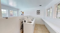 This shared laundry room is a practical, well-organized facility designed to serve residents with ease and efficiency. The space features clean white walls and light-toned tile flooring that contribute to a bright, sanitary environment. A row of top-load washing machines lines one wall, providing ample capacity for laundry needs. A dedicated folding counter extends along the adjacent wall, offering a convenient surface for sorting and folding clothes. Natural light filters in through windows on either side of the room, keeping the space feeling open and well-lit. Ventilation is provided via a ceiling vent, ensuring proper airflow. A bulletin board near the entrance serves as an informational hub, with posted guidelines and notices for users. The room is thoughtfully laid out, with clear organization and sufficient space to move comfortably between machines and the folding area. Overall, the laundry room is a clean, functional, and accessible shared amenity that prioritizes user convenience.