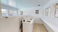 This shared laundry room is a practical, well-organized facility designed to serve residents with ease and efficiency. The space features clean white walls and light-toned tile flooring that contribute to a bright, sanitary environment. A row of top-load washing machines lines one wall, providing ample capacity for laundry needs. A dedicated folding counter extends along the adjacent wall, offering a convenient surface for sorting and folding clothes. Natural light filters in through windows on either side of the room, keeping the space feeling open and well-lit. Ventilation is provided via a ceiling vent, ensuring proper airflow. A bulletin board near the entrance serves as an informational hub, with posted guidelines and notices for users. The room is thoughtfully laid out, with clear organization and sufficient space to move comfortably between machines and the folding area. Overall, the laundry room is a clean, functional, and accessible shared amenity that prioritizes user convenience.