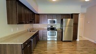 This two-story space offers a thoughtfully designed layout that balances functionality and style. The kitchen serves as a standout feature, showcasing rich, dark wood cabinetry that extends to the upper walls, providing generous storage throughout. Stainless steel appliances — including a refrigerator, electric range with oven, and over-the-range microwave — lend a modern, polished aesthetic. The countertops feature a neutral stone-look finish that complements the cabinetry, while a stainless steel sink with a sleek faucet adds to the kitchen's practical appeal. Warm hardwood-style flooring runs throughout, adding continuity and a welcoming feel to the space. The kitchen is laid out in an efficient configuration, maximizing workspace and flow. Soft, neutral wall tones enhance the brightness of the area, keeping the atmosphere open and inviting. The two-floor layout suggests additional living, sleeping, or utility spaces beyond the kitchen, offering versatility for a variety of uses. Overall, the space presents a clean, modern interior with durable finishes and a cohesive design that is both attractive and highly functional.