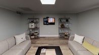 This single-floor space is designed as a versatile communal gathering area, thoughtfully arranged to accommodate a variety of activities within one open, flowing layout. Light wood-style flooring runs throughout, complementing the neutral gray-and-white color palette that gives the space a clean, contemporary feel.

The lounge area features a comfortable sectional sofa in soft gray, paired with accent pillows and a green upholstered chair. Framed black-and-white artwork and a wall-mounted coat rack add a welcoming, finished touch. Across the room, a dedicated media zone includes a wall-mounted flat-screen TV flanked by two open metal shelving units displaying decorative objects, creating a cozy entertainment nook with its own seating.

A bold black kitchen island with bar stools serves as a central anchor point, dividing the lounge from the dining and refreshment area. Nearby, a round white dining table surrounded by modern black chairs provides a casual spot for meals or meetings. A white cabinetry station with a countertop coffee setup rounds out the functional amenities.

Large windows line the perimeter walls, flooding the space with natural light and offering views of surrounding greenery. Recessed and panel ceiling fixtures provide ample ambient lighting. The overall ambiance is modern, comfortable, and multi-functional.