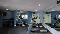 The fitness center at Tate's Creek Village is a dedicated, single-floor exercise space designed to support a well-rounded workout routine. The room features bold blue accent walls paired with a white ceiling, creating an energetic yet clean atmosphere conducive to exercise. Recessed lighting provides consistent illumination throughout the space, ensuring visibility across all workout areas.

The facility is equipped with a selection of cardio machines, including treadmills and elliptical trainers from Vision Fitness, positioned to allow comfortable use. A weight bench with associated equipment is also available for strength training. Large wall-mounted mirrors on multiple walls visually expand the room while allowing users to monitor their form during workouts.

Ventilation is provided via a ceiling-mounted HVAC unit, keeping the environment comfortable during exercise sessions. A water cooler is conveniently positioned within the room, offering easy hydration access. The flooring appears to be a dark, durable surface appropriate for a fitness environment. Natural light supplements the recessed lighting through at least one window visible in the mirror reflection, adding brightness to the overall space.