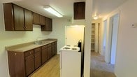 This single-floor residence offers a well-organized and functional layout designed for comfortable everyday living. The kitchen is a central feature of the space, outfitted with dark wood cabinetry that provides generous storage both above and below the countertops. A double-basin sink is positioned along the main wall, accompanied by a white electric range with coil burners and a refrigerator. Overhead fluorescent lighting illuminates the kitchen workspace efficiently. The kitchen opens toward a hallway that connects the living areas seamlessly, with light-toned carpet flowing into the adjacent rooms, creating a cohesive and warm atmosphere. A notable feature visible from the kitchen is a spacious walk-in storage closet with open shelving, offering significant organizational capacity. The ceilings are standard height and the overall color palette is neutral, with light walls complementing the darker cabinetry. Flush-mount ceiling fixtures provide consistent lighting throughout the common areas. The flooring transitions from wood-look vinyl in the kitchen to soft carpeting in the hallway and living spaces, adding comfort underfoot. The layout is compact yet efficient, making practical use of every area within the single-floor footprint.