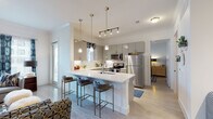 This single-floor residence showcases a thoughtfully designed open-concept layout that seamlessly connects the kitchen, dining, and living areas. The kitchen is a standout feature, outfitted with sleek gray cabinetry, white quartz countertops, and a full suite of stainless steel appliances including a refrigerator, range, and microwave. A large peninsula with an integrated sink and bar seating for multiple guests serves as the social centerpiece of the space. Pendant lighting in brushed gold tones and a track lighting system illuminate the area, while light wood-style flooring runs throughout, contributing to a clean, modern aesthetic. Natural light filters in through multiple windows dressed with stylish patterned curtains. The living area exudes a curated, design-forward character, featuring bold geometric rugs, layered seating, and eclectic accent pieces including a large indoor tree, textured cabinetry, and gold accents. The bedroom offers a calm retreat with window views, neutral walls, and coordinated décor. The bathroom features gray cabinetry, white quartz surfaces, a large mirror, and modern fixtures, maintaining the cohesive contemporary style found throughout the residence.