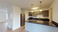 This single-floor residence offers a thoughtfully designed layout that balances functionality with modern style. The kitchen serves as a central highlight, featuring dark granite countertops, taupe cabinetry with brushed hardware, stainless steel appliances including a refrigerator, range, and microwave, and a peninsula with pendant lighting overhead. Light hardwood-style flooring flows throughout the main living areas, complementing the bright, white walls that lend a clean and airy feel to the space. The expansive living area is anchored by a wood-burning fireplace and benefits from natural light streaming in through a large window and sliding glass door that opens to an outdoor area. A stairwell with an arched window and pendant light connects to additional levels of the building. The space includes multiple bathrooms finished with light tile flooring, white fixtures, and simple, understated detailing. A dedicated in-unit laundry room houses a full-sized GE washer and dryer with built-in shelving above. Wide hallways and six-panel interior doors contribute to the open, well-connected flow of the floor plan. Overall, the space presents a comfortable and practical living environment with modern amenities throughout.