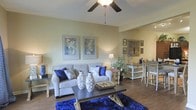 This single-floor residence offers a thoughtfully designed living space that blends comfort with decorative flair. The living room serves as the centerpiece, featuring warm beige walls that provide a neutral backdrop for a carefully curated interior. A ceiling fan with a light fixture anchors the room from above, contributing to both air circulation and ambient lighting.

The living area is furnished with a plush grey sofa accented by a rich combination of royal blue and patterned throw pillows, creating a bold contrast against the neutral tones of the room. A rustic wooden coffee table sits atop a striking royal blue area rug, adding a vibrant focal point to the space. Decorative ceramic vases and a small tray accessorize the table, while two framed botanical artworks adorn the wall above the sofa.

Hardwood-style flooring flows throughout, lending warmth and continuity to the space. Toward the rear, a transitional dining area is visible, featuring a wooden bar-height table paired with grey ladder-back chairs. A console table with a small lamp and a potted plant adds a welcoming touch between the two areas. The overall ambiance is warm, stylish, and inviting.