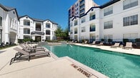 Located at 777 Dunlavy St in Houston's desirable 77019 zip code, this outdoor amenity space is designed to deliver a resort-like experience within a vibrant residential community. The centerpiece is a generously sized swimming pool with clear, blue-green water, framed by a broad, light-toned concrete sun deck lined with multiple lounge chairs — ideal for relaxing under the Texas sun. Adjacent to the pool sits an elevated spa with stone tile detailing, offering a dedicated space for relaxation. The surrounding architecture features a striking white-painted, multi-story building with arched detailing, dark accent trim, and large glass windows that bring an elegant, classic aesthetic to the outdoor environment. A clubhouse-style structure with floor-to-ceiling windows overlooks the pool area, seamlessly connecting the indoor and outdoor spaces. Lush green landscaping, manicured hedges, and mature trees provide shade and a park-like atmosphere throughout the grounds. A standout feature is a freestanding hammock set on a well-maintained grass lawn, adding a leisure-focused retreat within the community. Additional seating areas with outdoor sofas and shade umbrellas offer comfortable lounging spots. The entire area is enclosed by decorative iron fencing, ensuring a secure and private atmosphere.