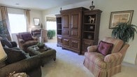 This single-floor residence exudes a classic, traditional charm throughout its well-appointed living spaces. The main living area serves as the heart of the home, anchored by a substantial dark wood armoire-style entertainment unit with paneled cabinet doors and open display shelving — an elegant focal point that adds both function and character to the room. Plush, neutral-toned carpeting runs throughout, contributing to the warm and comfortable ambiance. Seating is arranged thoughtfully, with a tufted striped armchair in muted rose tones and an olive-green upholstered ottoman adding layers of texture and color. A decorative tray with accent pieces rests on the ottoman, reinforcing the curated, lived-in feel of the space. An open passageway leads to an adjacent room, where natural light filters in through a window dressed with a tailored valance, illuminating a lush potted plant and hinting at additional living or dining space beyond. Subtle decorative touches — including framed artwork, a classic table lamp, and ornamental figurines displayed on the shelving unit — complete the traditionally styled interior, creating a cohesive and inviting atmosphere throughout.