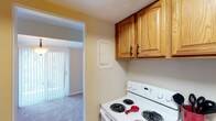 This single-floor residence offers a practical and straightforward layout across several well-defined spaces. The kitchen is equipped with a white coil-burner electric range and warm oak upper cabinetry featuring raised-panel doors and black hardware. Light-toned walls complement the functional design, and a doorway connects the kitchen directly to the main living area, which features vertical blinds and access to natural light through a sliding door.

The living area is generously sized, finished with neutral wall-to-wall carpeting and crisp white walls, providing a versatile, open canvas. Additional carpeted rooms serve as bedrooms, each featuring neutral-toned walls and windows with blinds that allow natural light to filter in. One bedroom includes a double set of bifold closet doors, offering substantial built-in storage with hanging rods. A second closet with bifold doors is also visible nearby, adding further storage convenience.

The bathroom features slate-look tile flooring, a vanity with warm wood cabinetry, a toilet, and a shower-tub combination enclosed by a full-length curtain rod. Overhead ventilation and a ceiling light fixture complete the space. Overall, the unit maintains a clean, neutral aesthetic throughout.