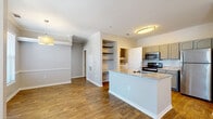 This single-floor residence offers a cohesive and functional layout designed with quality finishes throughout. Rich hardwood-style flooring flows across the main living areas, complemented by crisp white crown molding and clean baseboards that add a polished architectural touch. The living and dining areas are generously sized, each equipped with ceiling fans featuring integrated lighting, and brightened by windows with horizontal blinds that allow for natural light while maintaining privacy. The open-concept kitchen serves as a central hub, showcasing granite-topped countertops, a peninsula with a built-in sink, gray cabinetry with arched detailing, a subway tile backsplash, and a full suite of stainless steel appliances including a refrigerator, electric range, and microwave. Built-in open shelving adjacent to the kitchen provides convenient additional storage. The bathroom continues the refined aesthetic with gray cabinetry, a granite vanity top, a well-lit mirror, and hardwood-style flooring. A carpeted room offers a softer, quieter space suitable for a bedroom. Throughout the home, neutral wall tones and consistent finishes create a calm, cohesive ambiance that is both inviting and move-in ready.