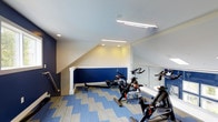 Located at 6060 Rossi Drive in Canal Winchester, Ohio, this dedicated fitness center offers a well-appointed single-floor space designed to support a variety of cardiovascular workouts. The room features a striking two-tone color scheme, with bold navy blue accent walls paired with crisp white upper walls and trim, creating an energizing yet clean aesthetic. Patterned carpet tiles in coordinating shades of blue and gray cover the floor, adding both comfort underfoot and visual interest to the space. The fitness area is equipped with multiple stationary cycling bikes and a rowing machine, providing residents or users with focused cardio training options. A floor-to-ceiling mirror along one wall expands the sense of space and allows users to monitor their form during workouts. Overhead, a combination of recessed lighting and linear fluorescent fixtures ensures the room is well-lit throughout. The angled ceiling line adds architectural character, following the natural roofline and giving the space a dynamic, loft-like quality. A staircase with a handrail is accessible from within the room, connecting the fitness area to the broader facility. Overall, the space is thoughtfully designed to be both functional and visually motivating.