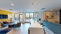 This single-floor communal space is thoughtfully designed to foster both social interaction and relaxed gathering. The open-plan layout flows seamlessly from one zone to the next, with large floor-to-ceiling windows along one wall flooding the interior with natural light and offering clear views of an adjacent outdoor terrace, complete with seating and greenery.

The main lounge area features multiple distinct seating arrangements, including a substantial blue sofa anchored by a patterned area rug, plaid accent chairs, and a rounded ottoman, creating an inviting, well-curated atmosphere. A bold teal accent wall serves as a focal point, housing a wall-mounted television, a geometric wooden shelving unit, and decorative artwork.

A long white countertop island with pendant lighting above acts as a central divider, doubling as a gathering or work surface with bar-height stools along one side. Adjacent to this is a well-appointed kitchenette featuring warm walnut cabinetry, open wooden display shelving, a quartz countertop, an undermount sink, and stainless steel appliances.

Large-format tile flooring runs throughout, lending a clean, contemporary feel. Recessed and pendant lighting complement the natural light, while curated artwork and decorative accessories complete the polished, modern aesthetic of the space.