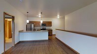 This two-floor space showcases a thoughtfully designed open-concept layout, with the kitchen serving as a central highlight. The kitchen is equipped with stainless steel appliances, including a refrigerator, electric range, and built-in microwave, all complemented by warm medium-toned wood cabinetry. A spacious central island with a granite-style countertop and an integrated sink provides both functional workspace and a natural gathering point. Pendant lighting fixtures hang above the island, adding both task lighting and a decorative element to the space. The flooring throughout features hardwood-style planks in a rich, warm tone, lending a cohesive and inviting feel. The walls are finished in a neutral white, keeping the space bright and versatile. A staircase landing is visible to the right, indicating the transition between the two floors and offering a sense of vertical openness. The overall ambiance is clean, modern, and functional, with a practical design that balances everyday usability with aesthetic appeal.