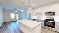 This thoughtfully designed single-story residence in Jacksonville, FL offers a clean, modern aesthetic throughout its well-appointed layout. The open-concept living area serves as the heart of the home, where the kitchen, dining, and living spaces flow seamlessly together beneath recessed lighting and ceiling fans. The kitchen is a standout feature, showcasing white shaker cabinetry, gray lower cabinets, a quartz waterfall island with pendant lighting, stainless steel appliances including a refrigerator with ice maker, dishwasher, range, and microwave, plus a classic subway tile backsplash. Durable hardwood-style flooring runs throughout the main living areas, complementing the bright, neutral color palette. The spacious bedroom is finished with plush carpet, a ceiling fan, and large windows that invite natural light and outdoor views. Two walk-in closets provide generous storage. The two bathrooms feature clean white finishes, quartz vanities, and a soaking tub. A dedicated laundry room houses a full-size washer and dryer. The living area opens to an outdoor-facing door, extending the sense of space and connection to the surroundings.