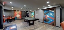 Located at 175 Forest Avenue in Hawthorne, NJ, this single-floor community clubhouse is thoughtfully designed to offer residents a variety of social and recreational spaces. The welcoming entrance lobby features tile flooring, recessed lighting, comfortable seating, a wall-mounted television, and a community bulletin board, creating an inviting first impression. Adjacent to the entry is a relaxed lounge area anchored by a large sectional sofa and accent chairs arranged around a flat-screen TV, set against a striking textured feature wall that adds visual depth and character to the room. Natural light filters in through windows dressed with colorful striped valances, a design detail that carries throughout the space and ties the areas together cohesively. A high-top dining area with bar-height tables and upholstered stools sits beside large windows overlooking the surrounding property, offering a bright spot for casual dining or conversation. The standout feature of the clubhouse is the expansive game room, complete with a full-size pool table illuminated by a pendant light, vibrant billiards-themed wall art, and a round poker table surrounded by orange upholstered chairs. Wood-look flooring throughout the game room adds warmth, while recessed lighting ensures the space is well-lit for entertainment. Together, these areas make for a versatile and engaging community gathering space.