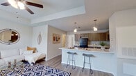 This single-story unit offers an efficient and thoughtfully designed layout across a comfortable footprint. The open-concept living area seamlessly connects to the kitchen, which is appointed with warm wood-toned cabinetry, black appliances including a refrigerator and range, light quartz-style countertops, and a peninsula with bar seating illuminated by pendant lighting overhead. Light wood-look flooring runs throughout, creating a cohesive aesthetic across the living, dining, and kitchen areas.

The spacious primary bedroom features a ceiling fan, a large window providing natural light, and a private door leading directly to an outdoor patio area. A generous walk-in closet with multi-level shelving and hanging rods offers substantial storage. The full bathroom is outfitted with a tub-shower combination, wood-toned upper cabinetry, a vanity with a white countertop, and a large mirror, maintaining the consistent warm-toned design language found throughout the unit.

Neutral gray walls, clean white ceilings, and modern hardware and fixtures contribute to a contemporary and cohesive interior. The layout maximizes functionality while maintaining an open, airy feel suitable for comfortable everyday living.
