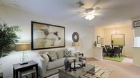 This single-floor residence offers a thoughtfully designed open-concept layout that promotes a natural flow between living and dining areas. The living room serves as the centerpiece of the home, featuring smooth white walls adorned with a large, bold framed artwork that adds a striking visual focal point. A ceiling fan with integrated lighting provides both comfort and illumination, while a decorative circular wall accent adds a modern touch near the entryway. The space is furnished with a plush gray sofa accented by vibrant patterned throw pillows, paired with a sleek metallic coffee table atop a textured area rug, creating a balanced blend of comfort and contemporary style. Tasteful accent lighting from a table lamp contributes to a warm and inviting ambiance. A white paneled door leads to the entry foyer, which connects seamlessly to the dining area visible in the background. The dining space features dark furniture set against green carpeting, offering a distinct and cozy contrast to the light-toned living area. Hardwood-style flooring in transitional areas adds warmth and continuity throughout the home. The overall design aesthetic is modern and well-curated, with attention to detail evident in every corner of this single-level residence.