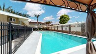 Situated along E Chapman Avenue in Orange, California, this single-story property offers a welcoming outdoor environment designed for relaxation and leisure. The centerpiece of the exterior is a generously sized rectangular swimming pool with clear, blue-green water, surrounded by a clean concrete pool deck. Tall palm trees and mature shade trees frame the pool area, lending a resort-like atmosphere under the Southern California sun. A wrought iron fence with decorative finials encloses the pool area, providing both security and an elegant aesthetic. Adjacent to the pool, a covered gazebo serves as a shaded retreat, featuring a warm canvas canopy with ornate ironwork detailing. The gazebo is furnished with wicker lounge chairs and curtain panels, creating a comfortable space for outdoor seating. Stone-accented pillars anchor the structure and complement the surrounding white lattice fencing and low stone walls that define the property boundaries. Lush green landscaping, including manicured shrubs and flowering plants, adds color and vibrancy throughout the grounds. The overall ambiance is bright, open, and inviting, reflecting the characteristic warmth of Orange County's outdoor living lifestyle.