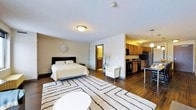 This single-floor studio offers a thoughtfully designed open-concept layout that seamlessly integrates living, sleeping, and dining functions into one cohesive space. Rich dark hardwood flooring runs throughout, anchoring the room with warmth and a sense of continuity.

The sleeping area is generously sized to accommodate a full bed and is accented by a bold dark headboard, complemented by neutral wall tones that keep the space feeling bright and airy. A geometric area rug defines the central living zone, where a white accent chair provides a comfortable seating option.

The kitchen occupies a partially separated alcove, distinguished by dark wood cabinetry that extends to the ceiling, a black refrigerator, and a black range. Pendant lighting above a small dining counter adds a modern finishing touch and enhances the kitchen's functionality. Flush-mount ceiling fixtures provide consistent ambient lighting throughout the main living area.

A doorway off the sleeping area leads to a private bathroom, offering a degree of separation and privacy within the open floor plan. The overall ambiance is cozy yet functional, with a neutral color palette, warm lighting, and practical finishes that make the space both inviting and highly livable.