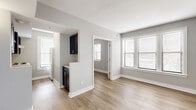 This 2-bedroom, 1-bathroom residence is thoughtfully laid out on a single floor, offering a clean, modern aesthetic with a cohesive neutral color palette of soft grays and crisp white trim throughout. Light wood-style flooring runs continuously across all rooms, lending warmth and visual continuity to the space.

The living room is bright and open, featuring multiple windows dressed with white horizontal blinds that allow generous natural light to fill the room. The adjacent kitchen is efficiently designed with dark navy cabinetry, light countertops, and white appliances, partially separated from the living area by a half-wall partition that maintains an open feel.

The primary bedroom is a comfortable, well-proportioned room with a distinctive arched window alcove — a notable architectural detail that adds character and charm. Both bedrooms feature large windows with outdoor views and ample wall space. A dedicated closet provides additional storage.

The full bathroom includes a white tiled tub-and-shower combination with chrome fixtures, maintaining the clean and functional aesthetic found throughout the home.

Overall, this residence balances practicality with a bright, airy ambiance, making it a well-appointed space with consistent finishes and a welcoming atmosphere from room to room.