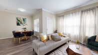 This thoughtfully designed single-floor residence offers a warm and contemporary living experience, highlighted by wood-look flooring, crown molding, and a neutral color palette that flows seamlessly throughout. The living room serves as the heart of the home, featuring large windows dressed with sheer curtains that invite abundant natural light. A geometric wooden coffee table and a mix of plush seating, including a deep green velvet accent chair, lend the space an elegant yet inviting character. Adjacent to the living area, the dining space accommodates a round wooden table with seating for four, accented by vibrant wall art that adds a bold focal point. The kitchen is well-appointed with white shaker-style cabinetry, granite-style countertops, a mosaic tile backsplash, stainless steel appliances, and a peninsula with bar seating beneath pendant lighting — ideal for casual dining or entertaining. The bedroom offers a tranquil retreat with a ceiling fan, large windows with sheer draping, and warm wood flooring. The bathroom is finished with a white vanity, granite-topped counters, a soaking tub with shower, and consistent wood-look flooring. Every room reflects a cohesive design sensibility that balances modern style with everyday comfort.