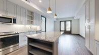 This contemporary single-floor residence offers a thoughtfully designed open-concept layout that seamlessly connects the kitchen and living areas. The kitchen is a standout feature, showcasing a large center island topped with granite countertops and accented by stylish gold pendant lights overhead. Light gray shaker-style cabinetry with under-cabinet lighting lines the walls, complemented by a white subway tile backsplash and a full suite of stainless steel appliances, including a French door refrigerator, microwave, and range. A built-in desk area with matching cabinetry adds a functional workspace within the kitchen. Rich hardwood flooring flows throughout the main living areas, while bold dark baseboards and dark-framed interior doors provide striking contrast against the neutral walls. The living area is bright and open, with French doors that invite natural light and offer access to the outdoors. The bedrooms feature plush carpeting, ceiling fans, and windows that allow for natural light. The full bathroom is elegantly finished with a granite-topped double vanity, dark cabinetry, large-format tile flooring, and a tiled shower-tub combination, completing this well-appointed residence.