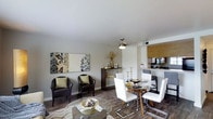 This well-appointed single-floor residence offers an inviting open-concept layout that seamlessly connects the living, dining, and kitchen areas. The living space is tastefully furnished with dark leather accent chairs adorned with sage green throw pillows, complemented by a round glass coffee table and a striking decorative wall canvas that adds an artistic focal point to the room. Rich wood-look flooring runs throughout, grounding the space with warmth and continuity. The dining area features a round wooden table surrounded by sleek white chairs with chrome legs, topped with elegant gold candle holders and decorative accents, all illuminated by a pendant light above. A dark wood sideboard provides additional storage and display space nearby. The kitchen, partially open to the dining area, is framed by warm wood cabinetry and includes modern appliances, making it both functional and visually connected to the rest of the living space. The neutral white walls and smooth ceilings create a bright, clean backdrop that allows the curated furnishings and gold accent tones to stand out. The overall ambiance is modern and refined, with a cohesive color palette of dark tones, white, and gold accents throughout.
