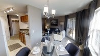 This single-floor residence offers a thoughtfully designed open-concept layout that seamlessly connects the kitchen, dining area, and living room into one cohesive and functional living space. The dining area anchors the layout with a glass-top table set beneath a brushed nickel chandelier, creating an elegant focal point that draws the eye upward. Woven placemats, decorative dishware, and a textured centerpiece vase add a refined, curated touch to the space. Adjacent to the dining area, the kitchen features warm wood cabinetry, a double basin sink, and tile flooring, offering a practical yet inviting workspace. The living room is finished with plush carpeting and a neutral, contemporary palette, furnished with a dark sofa accented with decorative pillows and a cream accent chair draped with a cozy throw. A framed cityscape artwork adds a sophisticated touch to the deep gray feature wall. A floor lamp provides warm ambient lighting, while sliding glass doors flood the space with natural light and offer access to an outdoor area, enhancing the sense of openness and connection to the exterior. The overall ambiance is modern, comfortable, and tastefully styled throughout.