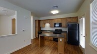 This single-floor space centers around a well-appointed kitchen designed for practical, everyday use. The kitchen features warm-toned wood cabinetry that extends to the upper walls, providing generous storage capacity. Dark granite-style countertops run along the perimeter and continue onto a peninsula, which offers additional workspace and casual dining potential.

The kitchen is equipped with black appliances, including a gas range, over-the-range microwave, dishwasher, and a full-sized refrigerator. A stainless steel sink is positioned along the back wall beneath the upper cabinetry, complemented by a dedicated ventilation vent overhead. The layout follows a functional galley-style arrangement along the back wall, enhanced by the extended peninsula that creates a natural division between the kitchen and the adjoining living area.

Hardwood-style flooring flows throughout the space, adding warmth and visual continuity. Overhead, flush-mounted lighting provides consistent illumination. The neutral wall tones complement the cabinetry and countertops, contributing to a cohesive, understated aesthetic throughout the space.