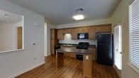 This single-floor space centers around a well-appointed kitchen designed for practical, everyday use. The kitchen features warm-toned wood cabinetry that extends to the upper walls, providing generous storage capacity. Dark granite-style countertops run along the perimeter and continue onto a peninsula, which offers additional workspace and casual dining potential.

The kitchen is equipped with black appliances, including a gas range, over-the-range microwave, dishwasher, and a full-sized refrigerator. A stainless steel sink is positioned along the back wall beneath the upper cabinetry, complemented by a dedicated ventilation vent overhead. The layout follows a functional galley-style arrangement along the back wall, enhanced by the extended peninsula that creates a natural division between the kitchen and the adjoining living area.

Hardwood-style flooring flows throughout the space, adding warmth and visual continuity. Overhead, flush-mounted lighting provides consistent illumination. The neutral wall tones complement the cabinetry and countertops, contributing to a cohesive, understated aesthetic throughout the space.