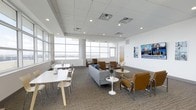 This single-floor commercial space offers a bright and professionally designed lounge environment ideal for collaborative or hospitality-focused use. The open-plan layout is anchored by large, multi-pane windows that stretch across the perimeter walls, flooding the interior with natural light and offering expansive outward views. The ceiling features a recessed tile grid with integrated HVAC vents and recessed lighting, contributing to a clean, polished aesthetic throughout.

The space is thoughtfully organized into distinct seating zones. A casual dining or meeting area near the windows includes white rectangular tables paired with modern white chairs, offering a crisp, minimal look. A central lounge zone features a gray sectional sofa, round side tables, and sleek tan leather chairs with chrome frames — creating a warm yet contemporary atmosphere. Additional accent chairs and small round tables are positioned to encourage intimate conversation or individual use.

Neutral tones dominate the design, with white walls, a patterned commercial-grade carpet in gray and taupe, and carefully curated decor including potted plants and floral arrangements. A wall-mounted display screen adds a functional media element. The overall ambiance is professional, welcoming, and well-appointed, suitable for a corporate lounge, waiting area, or executive suite.