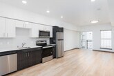 This contemporary single-floor unit offers a clean, modern design throughout, characterized by light hardwood flooring, crisp white walls, and recessed lighting that create a bright and welcoming atmosphere. The open-concept layout seamlessly connects the kitchen and living area, maximizing the sense of space and functionality.

The kitchen is thoughtfully appointed with a two-tone cabinetry design — white upper cabinets paired with rich dark wood lower cabinets — complemented by light quartz countertops and a subtle tile backsplash. Stainless steel appliances, including a refrigerator, stove, microwave, and dishwasher, round out this well-equipped space. A convenient in-unit laundry closet is accessible directly from the kitchen area.

The living area is generously sized and filled with natural light, courtesy of large windows and a sliding glass door that opens onto a private balcony. This outdoor access adds an airy, open quality to the living space and provides pleasant views beyond.

The bathroom features a clean, minimalist aesthetic with a wood-toned vanity, light countertop, a full-width mirror, and a combined tub and shower. Consistent with the rest of the unit, the design prioritizes simplicity and functionality. Overall, this space delivers a polished, move-in-ready environment with thoughtful modern finishes throughout.