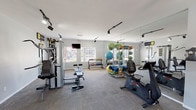 This fitness center offers a comprehensive workout environment designed to accommodate a wide range of exercise routines. The space is finished with neutral gray carpeting and light gray walls, creating a clean and focused atmosphere conducive to training. Track lighting runs along the ceiling, providing bright, directed illumination throughout the room. A wall of floor-to-ceiling mirrors on one side visually expands the space and allows users to monitor their form during workouts. Natural light filters in through windows along the exterior wall, adding to the open and energizing ambiance. The equipment selection is diverse and well-organized, featuring a multi-function cable and weight machine, an adjustable workout bench, a stationary exercise bike, and a stair climber. A dedicated free weights station includes a selection of kettlebells and dumbbells neatly arranged on a rack. A storage tower holds an assortment of stability and medicine balls in various sizes, along with foam rollers for stretching and recovery. A wall-mounted flat-screen television is positioned near the windows for entertainment during workouts. The single-floor layout ensures convenient, accessible use of all equipment in one streamlined space.