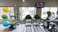 This fitness center is a thoughtfully designed workout space that provides residents with everything needed for a comprehensive exercise routine. The room is anchored by multiple treadmills positioned to take full advantage of expansive floor-to-ceiling windows, offering motivating views of a sparkling pool and tropical palm trees beyond. Natural light floods the space, creating an open and energizing atmosphere. A wall-mounted flat-screen television keeps members entertained during cardio sessions. To one side, a well-organized equipment rack holds an assortment of medicine balls in various weights alongside exercise mats and foam rollers, catering to stretching, core work, and functional fitness training. The flooring features a durable, wood-look surface that is both practical and visually clean. Recessed and track lighting supplements the ample natural light, ensuring the space remains bright at any hour. The neutral color palette of the walls and finishes contributes to a calm, focused environment. Overall, this fitness center balances practicality and comfort, offering a welcoming space designed to support a wide range of workout styles and fitness goals.