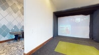 This single-floor facility is thoughtfully designed for golf enthusiasts, combining a high-tech simulator bay with a comfortable and visually striking lounge space. The simulator area features a Full Swing golf simulator set within a dark-trimmed enclosure, complete with a projection screen, a bright green hitting mat, and grey carpet flooring — creating a focused, immersive environment for practice and play. Wood baseboard trim adds a warm, finished touch throughout. Adjacent to the simulator is a generously sized lounge that exudes a contemporary yet inviting character. A bold geometric argyle-patterned accent wall in tones of grey, teal, and white serves as a dramatic focal point, complementing the patterned area rug underfoot. Seating includes a tufted black leather sofa, a coordinating leather bench, and plush burgundy accent chairs, all arranged to encourage relaxed conversation. Large windows allow abundant natural light to fill the lounge, offering views to the outside. A stand-up bar-height table and additional side tables complete the social atmosphere. The transition between the carpeted lounge and the grey-carpeted simulator bay is seamless, making the space ideal for groups who want to mix competitive play with comfortable downtime.