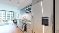 Located at 1059 Water St in Tampa's Water Street district, this single-floor residence offers a thoughtfully designed modern interior with a neutral palette and warm hardwood flooring throughout. The galley-style kitchen is efficiently organized, featuring gray-toned cabinetry that extends to the ceiling, a white subway tile backsplash, stainless steel appliances including a built-in microwave, wall oven, induction cooktop, and full-size refrigerator, all complemented by clean white countertops. The kitchen opens toward a dining area, creating a natural flow into the living space. The living room is a standout area, anchored by expansive floor-to-ceiling windows that frame urban views of the surrounding cityscape and flood the space with natural light. A warm-toned media console and woven accent chair add texture and character to the room. The bedroom continues the floor-to-ceiling window design, offering the same light-filled urban views, with warm wood flooring and a generous closet. The bathroom features large-format tile flooring, a wood-toned vanity with a light stone countertop, and minimalist hardware, contributing to the overall clean, contemporary aesthetic found throughout the space.