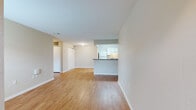 This single-floor unit is designed with a practical, open layout that connects the main living area to the kitchen, creating a cohesive and functional flow. Warm hardwood-style flooring runs consistently throughout the space, complementing the neutral beige and white wall tones that lend a clean, bright ambiance.

The living area is generously proportioned, offering ample room for various furniture arrangements. A front entry door flanked by a window allows natural light to filter in, enhancing the openness of the space. The kitchen features a peninsula with a dark countertop that separates it from the living area, providing both a visual anchor and additional workspace. White cabinetry lines the kitchen walls, accompanied by a full-size refrigerator, a range, and a sink — delivering all essential appliances in a compact, efficient galley-style configuration.

Additional rooms offer flexibility and are equally finished with hardwood-style flooring and windows that bring in natural light. The bathroom is well-appointed with a full soaking tub and shower combination, a white vanity with under-sink storage, a large mirror, and a bright vanity light fixture above. Clean white finishes and neutral tones carry throughout, maintaining a consistent and understated aesthetic across the entire unit.