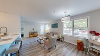 Located at 7102 Rock Ridge Lane in Alexandria, Virginia, this single-floor residence offers a thoughtfully designed layout with a cohesive, modern aesthetic throughout. The living area serves as the centerpiece of the home, featuring light-toned hardwood-style flooring, neutral gray walls, and an abundance of natural light streaming through multiple large windows fitted with white blinds. A stylish drum pendant light anchors the space, which is open and generously proportioned. The room flows seamlessly into a dedicated seating area near the entry, ideal for use as a home office or reading nook, also benefiting from natural light and direct access to the front door.

The kitchen is well-equipped with two-tone cabinetry—white upper cabinets and gray lower cabinets—paired with a light subway tile backsplash, white countertops, and stainless steel appliances including a range, microwave, and refrigerator. Light tile flooring completes the functional yet attractive space.

The bedroom is a serene retreat, featuring soft neutral tones, a mirrored sliding closet, and a sliding glass door that opens to a private outdoor area with greenery views. The bathroom maintains the home's clean design with white cabinetry, a tub-shower combination, and wood-style flooring.