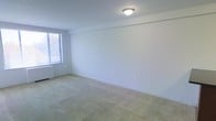 This single-floor space presents a clean, minimalist interior centered around a generously sized carpeted room. The room features light beige wall-to-wall carpeting that contributes to a neutral, understated aesthetic throughout. The walls are finished in a crisp white, lending an open and airy feel to the interior. A single window fitted with horizontal blinds allows natural light to filter into the space, brightening the room while offering privacy control. A flush-mount ceiling light fixture provides overhead illumination, complementing the natural light source. The room's simple layout offers considerable flexibility for furniture arrangement and use. A standard electrical outlet is visible along the lower wall, reflecting practical, functional detailing. The overall ambiance is quiet and straightforward, with a blank-canvas quality that makes the space highly adaptable to a variety of configurations and styles.
