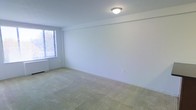 This single-floor space presents a clean, minimalist interior centered around a generously sized carpeted room. The room features light beige wall-to-wall carpeting that contributes to a neutral, understated aesthetic throughout. The walls are finished in a crisp white, lending an open and airy feel to the interior. A single window fitted with horizontal blinds allows natural light to filter into the space, brightening the room while offering privacy control. A flush-mount ceiling light fixture provides overhead illumination, complementing the natural light source. The room's simple layout offers considerable flexibility for furniture arrangement and use. A standard electrical outlet is visible along the lower wall, reflecting practical, functional detailing. The overall ambiance is quiet and straightforward, with a blank-canvas quality that makes the space highly adaptable to a variety of configurations and styles.