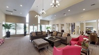 This single-floor communal space exudes a refined, welcoming atmosphere defined by dramatic vaulted ceilings that rise to impressive heights throughout the main lounge area. Dual elegant chandeliers cast warm, ambient light across the open-plan interior, complemented by recessed lighting and an abundance of natural light streaming through large windows along the perimeter walls.

The main lounge is thoughtfully furnished with plush taupe sofas, a tufted ottoman, and vibrant red accent chairs arranged around a dark wood coffee table, creating a comfortable and visually dynamic seating area. Rich hardwood flooring transitions to neutral carpet, delineating the various zones within the open layout.

A standout architectural feature is the expansive marble-surround fireplace, framed by a large decorative mirror and flanked by tall indoor tropical plants that introduce a lush, organic element to the space. A flat-screen television and curated wall art add to the lounge's refined character.

Adjacent to the main lounge, a more intimate seating nook provides a quieter retreat, while large interior windows offer clear views into a professional office area furnished with dark wood desks and workstations. Lush potted plants are positioned throughout, enhancing the space's warm and inviting ambiance.
